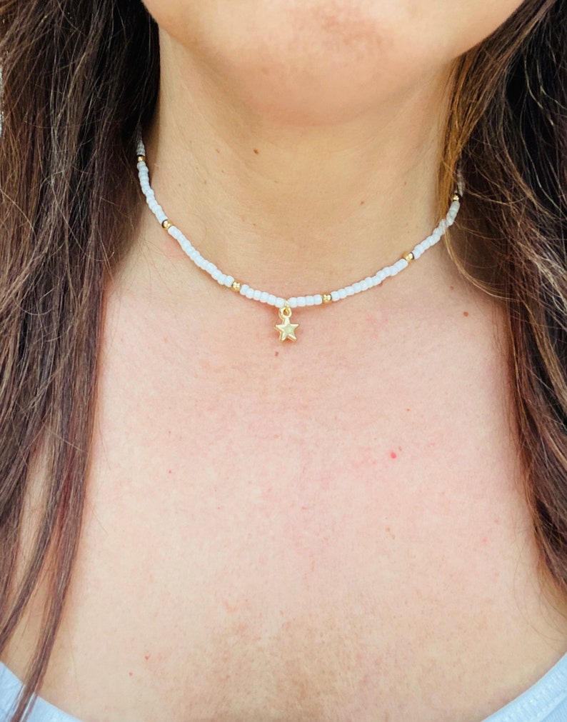 Pu&ograve; includere: Collana girocollo con perline bianche con dettagli dorati e un ciondolo a stella dorata. La collana &egrave; indossata intorno al collo. Le piccole perline distanziate creano un design semplice ed elegante. Un gioiello delicato.