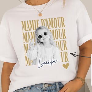 Peut inclure: Un t-shirt blanc à manches courtes avec un motif de texte doré répété Mamie D'Amour. Au centre, une photographie en noir et blanc d'un enfant portant des lunettes de soleil et faisant un signe de paix, avec le nom Louise et un cœur doré.