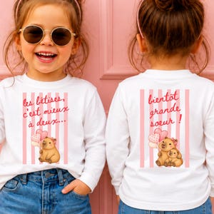 Peut inclure: Deux t-shirts blancs à manches longues avec des rayures verticales roses et un motif de capybara. Le t-shirt de gauche porte le texte 'les bêtises, c'est mieux à deux...' et celui de droite 'bientôt grande soeur!' avec des ballons en forme de cœur.