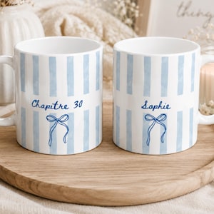 Peut inclure: Deux tasses en céramique blanche ornées de rayures verticales bleu clair. La tasse de gauche porte l'inscription "Chapitre 30" en écriture bleue au-dessus d'un nœud dessiné. La tasse de droite porte le prénom "Sophie" au-dessus d'un nœud.