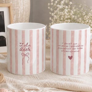 Peut inclure: Deux tasses en céramique blanche avec des rayures verticales roses. L'une porte l'inscription Tata Adèle avec un nœud, l'autre le texte il paraît que les amies formidables deviennent des tatas extraordinaires avec un petit cœur.