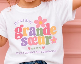 Op de afbeelding: Een wit T-shirt met korte mouwen en de tekst 'JE VAIS ÊTRE grande sœur EN 2027 ET ÇA SERA MOI QUI COMMANDE !' in kleurrijke, afgeronde letters, versierd met twee roze en gele bloemafbeeldingen en twee kleine roze hartjes.