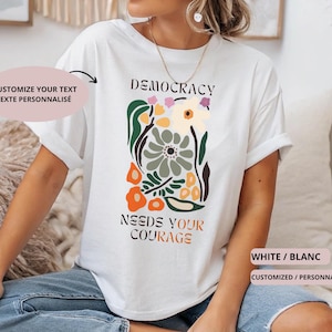 Può includere: T-shirt bianco con la scritta "DEMOCRACY NEEDS YOUR COURAGE" e un design floreale in verde, arancione e giallo. La maglietta ha anche la scritta "CUSTOMIZE YOUR TEXT TEXTE PERSONNALISÉ" e "WHITE / BLANC CUSTOMIZED / PERSONNALISÉ".
