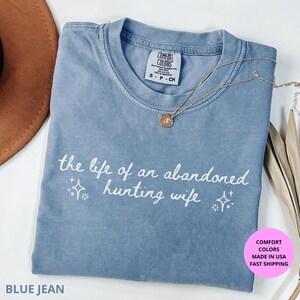 May include: Blue jean Comfort Colors t-shirt with the phrase "the life of an abandoned hunting wife" in white script. A gold necklace with a pendant rests on the shirt. The shirt is folded and displayed on a white surface.