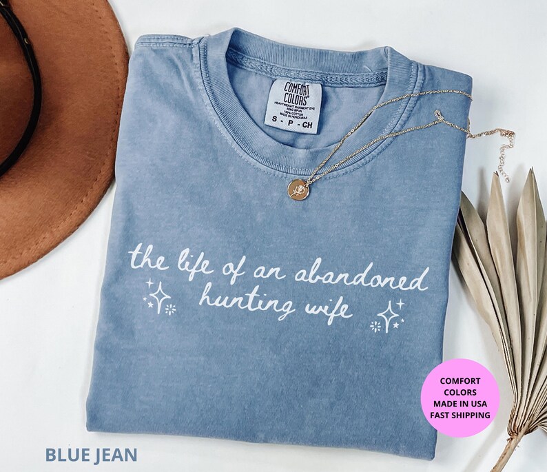 May include: Blue jean Comfort Colors t-shirt with the phrase "the life of an abandoned hunting wife" in white script. A gold necklace with a pendant rests on the shirt. The shirt is folded and displayed on a white surface.