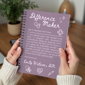 Könnte beinhalten: Ein lila Notizbuch mit Spiralbindung, auf dem die Worte "Difference Maker" stehen. Das Notizbuch enthält eine Definition einer Krankenschwester mit dem Namen "Emily Watson, RN" am unteren Rand. Das Notizbuch wird von einer Person gehalten.