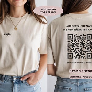 May include: Beige t-shirt with the word "single" printed on the front. The back of the shirt features a QR code and German text. The shirt also has the words "NATURAL" and "CUSTOMIZED" printed on it.