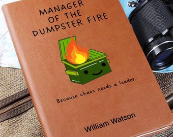 May include: A brown leather-bound journal with the text "Manager of the Dumpster Fire" and a cartoon image of a green dumpster with flames on top. The text "Because chaos needs a leader." is below the image. The name "William Watson" is printed at the bottom of the journal.