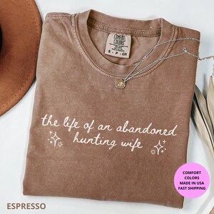 May include: Espresso-colored t-shirt with the text "the life of an abandoned hunting wife" in white script. The shirt has a crew neck and short sleeves. A silver necklace with a pendant is draped across the shirt.