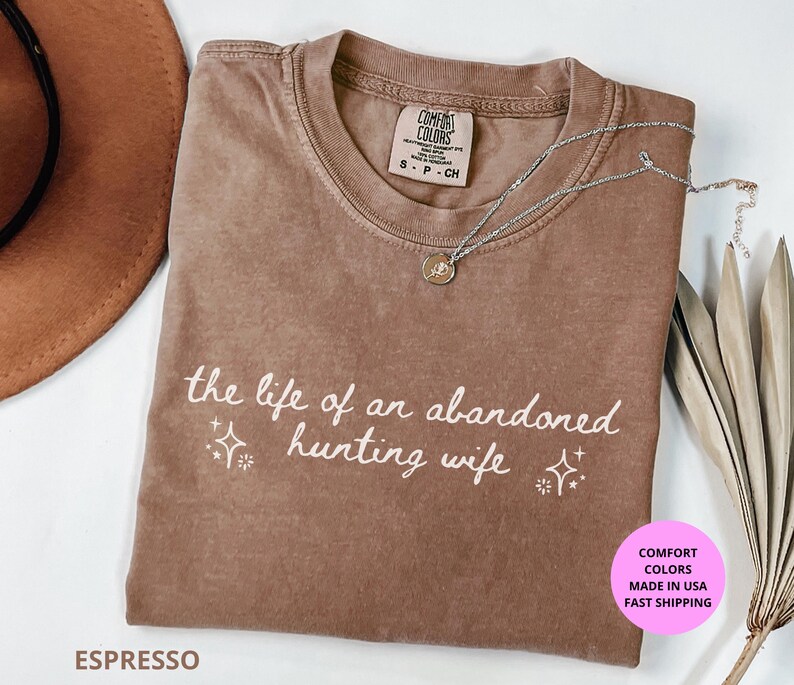 May include: Espresso-colored t-shirt with the text "the life of an abandoned hunting wife" in white script. The shirt has a crew neck and short sleeves. A silver necklace with a pendant is draped across the shirt.