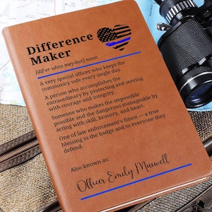 Peut inclure: Un carnet en cuir marron avec les mots "Difference Maker" et une définition imprimée sur la couverture. La couverture présente également un motif de cœur avec le drapeau américain et une ligne bleue. Le carnet est personnalisé avec le nom "Officer Emily Maxwell".