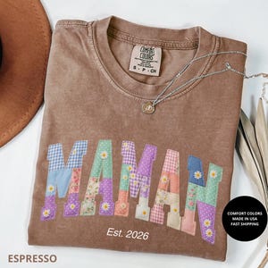 Peut inclure: T-shirt Comfort Colors couleur expresso avec le mot "MAMA" en lettres patchwork. Les lettres sont de différentes couleurs pastel et motifs, avec de petits accents de marguerites. Le texte "Est. 2026" est en dessous du mot "MAMA". Un collier argenté est sur le t-shirt.