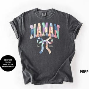 Peut inclure: Un t-shirt gris foncé avec le mot "MAMAN" en lettres colorées en patchwork. En dessous, un nœud papillon en patchwork avec l'année "2026". Le t-shirt a des manches retroussées et le mot "PEPPER" est imprimé à droite.