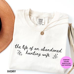 May include: Ivory-colored t-shirt with the text "the life of an abandoned hunting wife" in black script. The shirt has a crew neck and a Comfort Colors tag. A gold necklace and a brown hat are also visible.