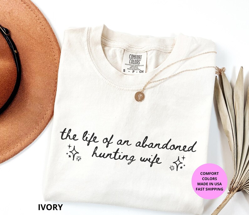 May include: Ivory-colored t-shirt with the text "the life of an abandoned hunting wife" in black script. The shirt has a crew neck and a Comfort Colors tag. A gold necklace and a brown hat are also visible.
