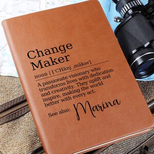 May include: A brown leather journal featuring the text "Change Maker" and its definition in black. The definition describes a visionary who transforms lives. The name "Marina" is also printed on the journal.