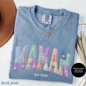 Peut inclure: T-shirt bleu jean Comfort Colors avec le mot "MAMA" en lettres patchwork. Chaque lettre est d'une couleur et d'un motif différents, avec de petits accents de marguerites. Un collier doré et un chapeau marron sont également visibles. Le texte "Est. 2026" est en dessous.