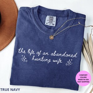 May include: A True Navy Comfort Colors t-shirt with the text "the life of an abandoned hunting wife" in white script. A gold necklace with a pendant rests on the shirt. The shirt is folded with a brown hat and dried plant in the background.
