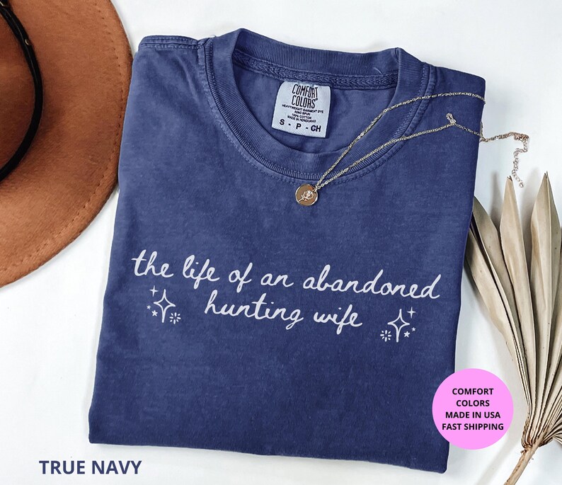 May include: A True Navy Comfort Colors t-shirt with the text "the life of an abandoned hunting wife" in white script. A gold necklace with a pendant rests on the shirt. The shirt is folded with a brown hat and dried plant in the background.
