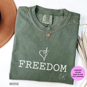 May include: A moss green t-shirt with the word "FREEDOM" printed in white. A heart and cross graphic is above the word. A gold necklace is draped across the shirt. The shirt has a Comfort Colors tag.