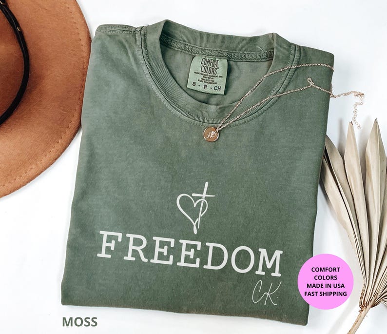 May include: A moss green t-shirt with the word "FREEDOM" printed in white. A heart and cross graphic is above the word. A gold necklace is draped across the shirt. The shirt has a Comfort Colors tag.