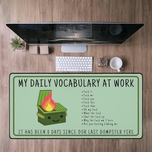Op de afbeelding: Groene bureaumat met de tekst "MY DAILY VOCABULARY AT WORK" en een lijst met krachttermen. Een cartoon vuilnisbak met vlammen staat in het midden. De tekst "IT HAS BEEN 0 DAYS SINCE OUR LAST DUMPSTER FIRE" staat onderaan.