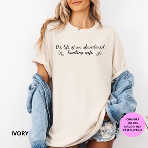 May include: Ivory-colored t-shirt with the text "the life of an abandoned hunting wife" in black script. The shirt has a relaxed fit and is paired with a denim jacket. The image also includes a pink circle with text.