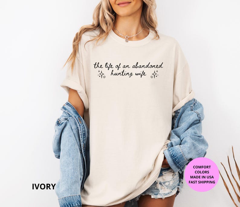May include: Ivory-colored t-shirt with the text "the life of an abandoned hunting wife" in black script. The shirt has a relaxed fit and is paired with a denim jacket. The image also includes a pink circle with text.