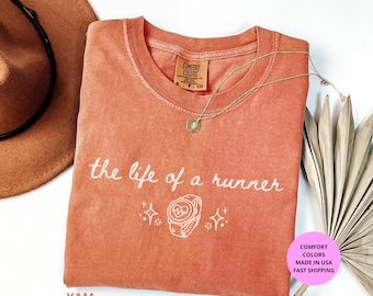 May include: A coral-colored t-shirt with the text "the life of a runner" in white script. The shirt features a graphic of a smartwatch and sparkles. A gold necklace and a brown hat are also visible.