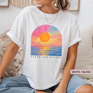 Może przedstawiać: Biała koszulka z kolorowym wzorem zachodu słońca w łuku, z czarnym napisem "TAXER LES RICHES". Projekt zawiera odcienie różu, pomarańczy, żółci i niebieskiego. To codzienne, swobodne ubranie.