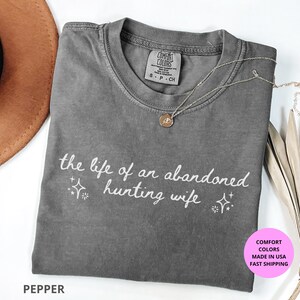 May include: Pepper-colored t-shirt with the text "the life of an abandoned hunting wife" in white script. The shirt has a crew neck and short sleeves. A gold necklace with a pendant is draped on the shirt.