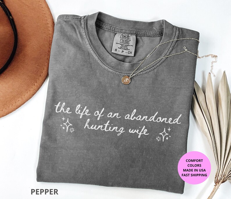 May include: Pepper-colored t-shirt with the text "the life of an abandoned hunting wife" in white script. The shirt has a crew neck and short sleeves. A gold necklace with a pendant is draped on the shirt.