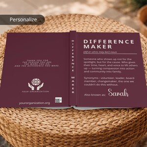Peut inclure: Un carnet bordeaux ouvert, affichant les mots "DIFFERENCE MAKER" en blanc. La définition de "Difference Maker" est imprimée sur la page de droite. La page de gauche contient un texte de remerciement et un logo d'ONG.