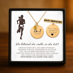May include: Gold-colored necklace with two round pendants, one with "26.2" and hearts, the other for custom engraving. The necklace is displayed in a black box with a runner silhouette and inspirational text. The text reads "She believed she could, so she did!"