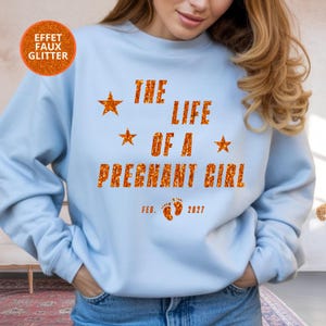 Può includere: Una felpa girocollo azzurra con testo arancione effetto glitter che recita THE LIFE OF A PREGNANT GIRL, accompagnata da tre stelle arancioni e dal testo FEB. 2027 con due piccole impronte di piedi di neonato arancioni al centro.
