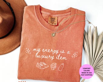 May include: A coral-colored t-shirt with the phrase "my energy is a luxury item" in white script. The shirt features white line drawings of sneakers, a watch, and a water bottle. A gold necklace and a brown hat are also visible.
