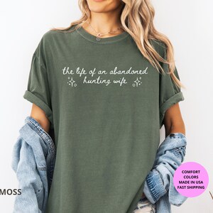 May include: Moss green t-shirt with the text "the life of an abandoned hunting wife" in white script. The shirt has short sleeves and a relaxed fit. The shirt is made of a soft material.