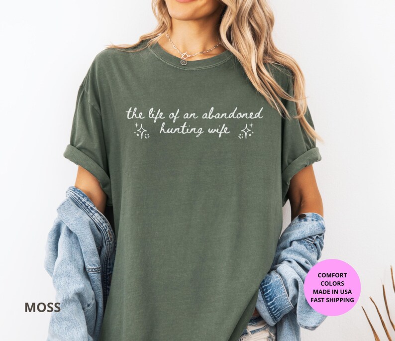 May include: Moss green t-shirt with the text "the life of an abandoned hunting wife" in white script. The shirt has short sleeves and a relaxed fit. The shirt is made of a soft material.