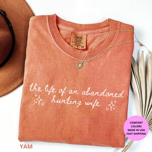 May include: A coral-colored t-shirt with the text "the life of an abandoned hunting wife" in white script. A gold necklace with a pendant rests on the shirt. A brown hat and dried palm leaves are in the background.