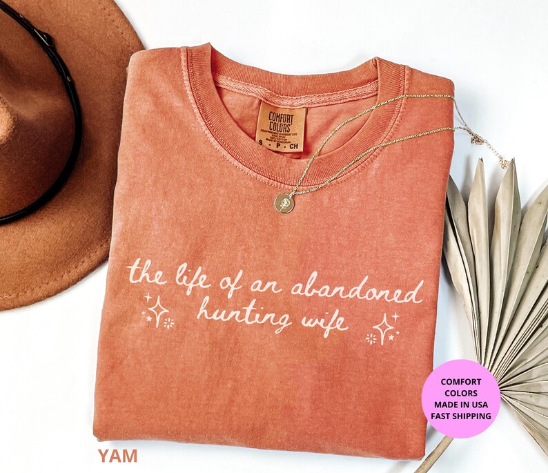 May include: A coral-colored t-shirt with the text "the life of an abandoned hunting wife" in white script. A gold necklace with a pendant rests on the shirt. A brown hat and dried palm leaves are in the background.