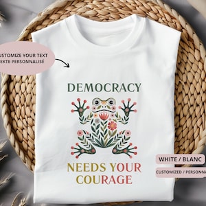Peut inclure: T-shirt blanc avec l'inscription "DEMOCRACY NEEDS YOUR COURAGE" en vert et rouge. Une illustration de grenouille stylisée avec des détails floraux est centrée sur le t-shirt. Le t-shirt est présenté sur un panier tressé.