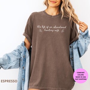 May include: Espresso-colored t-shirt with the text "the life of an abandoned hunting wife" in a script font. The shirt is worn with a light blue denim jacket. The image also includes a pink circle with text.