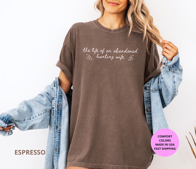 May include: Espresso-colored t-shirt with the text "the life of an abandoned hunting wife" in a script font. The shirt is worn with a light blue denim jacket. The image also includes a pink circle with text.