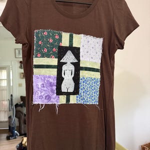 May include: A brown short-sleeve t-shirt featuring a central square patch made of various floral and solid fabric pieces. The patch includes a black rectangular inset with a white line-art illustration of a figure wearing a mushroom-shaped hat.