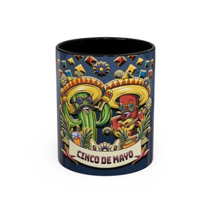 Puede incluir: Una taza de cerámica negra con una ilustración colorida de dos personajes de dibujos animados que llevan sombreros y tocan instrumentos musicales. El texto "Cinco de Mayo" está impreso en la taza.