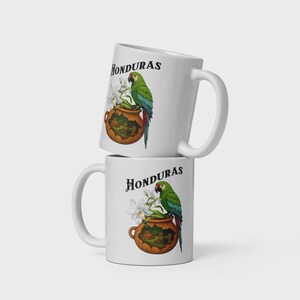 Puede incluir: Dos tazas de cerámica blanca apiladas. Cada taza presenta la palabra "Honduras" en escritura negra, un loro verde, una olla marrón y flores blancas. El loro tiene alas azules y cola roja.