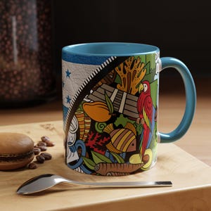 Puede incluir: Una taza de café de cerámica azul con un diseño colorido que presenta un loro, un pez y plantas tropicales. La taza tiene un borde y un asa blancos.