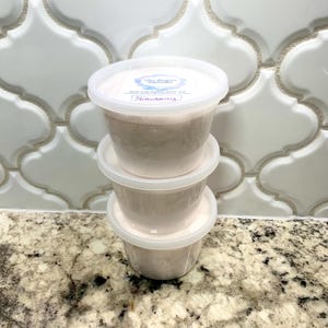 May include: Three clear plastic containers stacked on top of each other. Each container is filled with a light pink, fluffy substance. The top container has a white label with the text "The Sugar Mama" and "Strawberry".