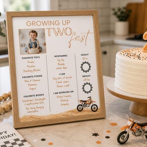 Op de afbeelding: Een ingelijst verjaardagsbord met de tekst "Growing Up Two Fast" en een foto. Het bord bevat een lijst met favoriet speelgoed, eten en boeken. Een witte taart met een oranje nummer twee kaars en een speelgoedmotorfiets zijn ook zichtbaar.