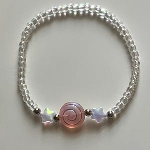 May include: A beaded bracelet featuring a central pink spiral charm flanked by two iridescent star-shaped beads and two small silver-toned metallic spheres, all strung on a cord with numerous clear, iridescent, round glass seed beads.